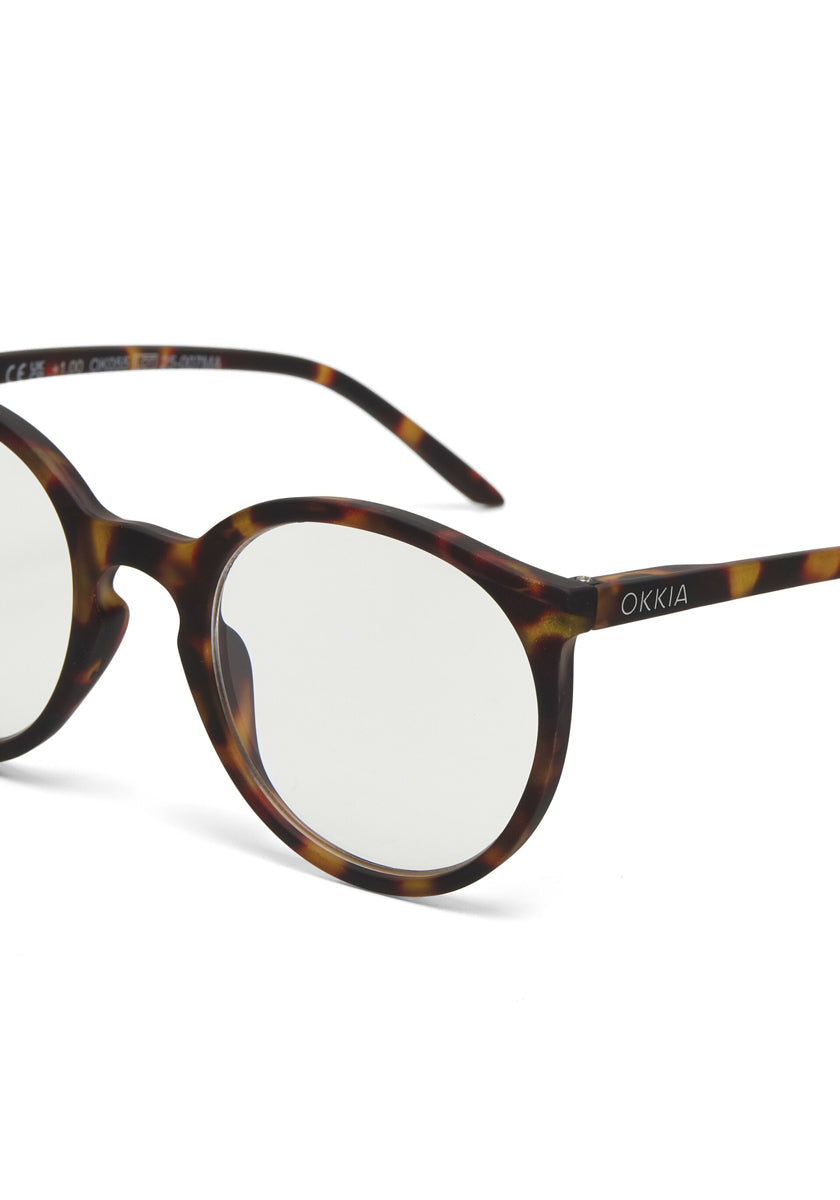 Close-up of Okkia tortoiseshell round eyeglasses with clear lenses in a classic circular design. The frame features a unique Schildpattmuster, and &quot;Okkia&quot; appears in white on the left arm. The background is simple and white.