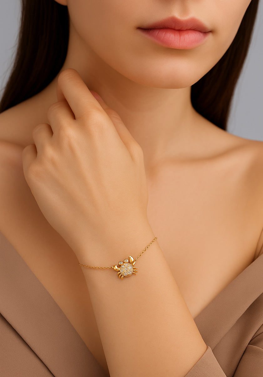 A woman with straight brown hair wears a taupe off-shoulder top by Princess Goes Hollywood and a gold-plated crab charm bracelet adorned with sparkling stones, her hand gently highlighting the jewelry against her light skin.