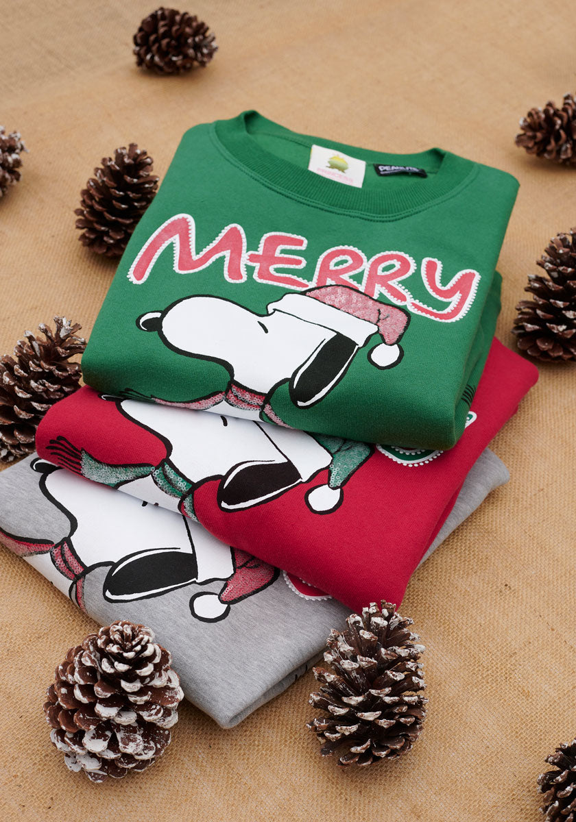 Drei gefaltete Princess Goes Hollywood Snoopy-Pullover liegen auf Sackleinen mit Tannenzapfen und Kunstschnee: ein grüner Merry Christmas-Pullover mit Snoopy in einer Weihnachtsmann-Mütze sowie ein roter und grauer Damen Weihnachtspullover mit festlichen Snoopy-Motiven.