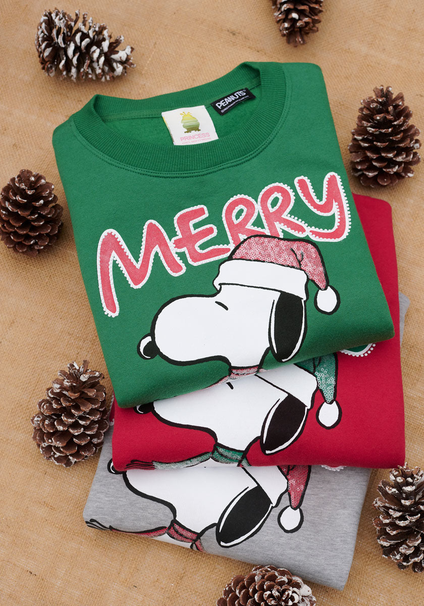 Drei gefaltete Princess Goes Hollywood Snoopy Weihnachtspullover - grün, rot und grau - sind auf Sackleinen mit schneebestäubten Tannenzapfen gestapelt. Der oberste grüne Pullover zeigt Snoopy mit einer Weihnachtsmannmütze über dem Wort "MERRY" in festlichen Buchstaben.