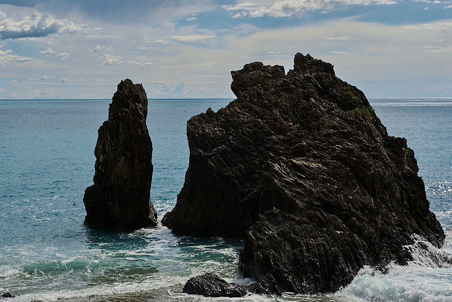 Two jagged, dark rocks stand in turquoise ocean water, with waves gently crashing against them. The sky above is partly cloudy, with sunlight breaking through, casting subtle reflections on the seas surface.