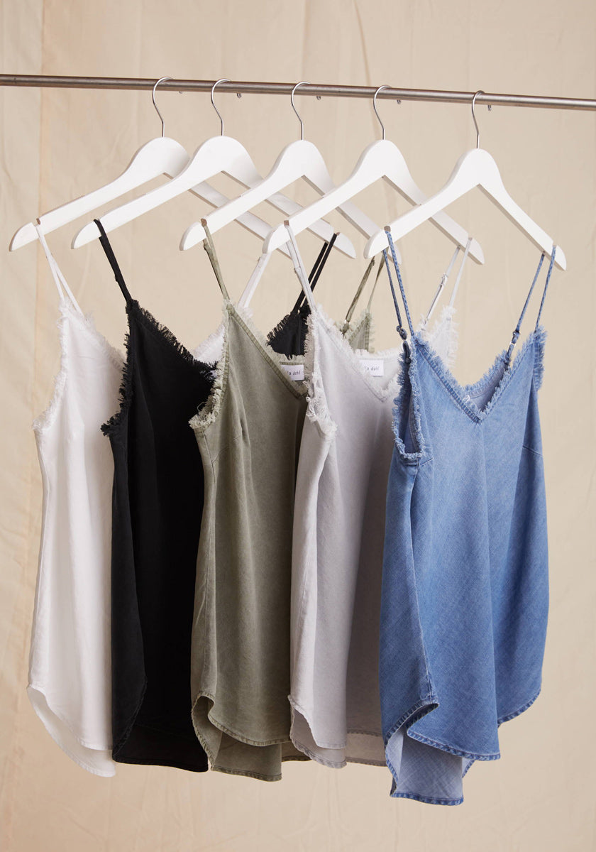 Five Bella Dahl sleeveless tops—denimoptik, breathable Lyocell, and more—hang on white hangers in white, black, olive green, light gray, and denim blue against a beige fabric background.