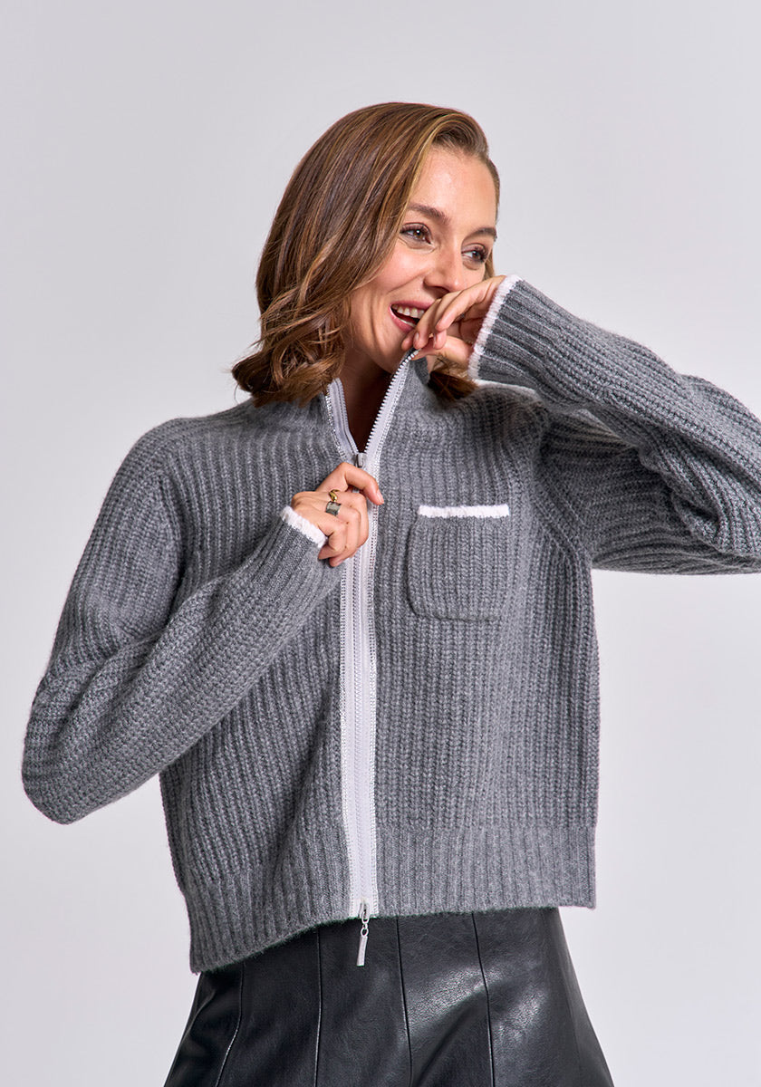 A woman with wavy brown hair smiles in a ribbed gray Strickjacke by Princess Goes Hollywood featuring a white zipper and front pocket, holding the collar near her face. She pairs it with a black leather skirt against a plain light gray background.