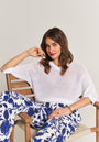 A woman sits on a wooden chair, wearing a Princess Goes Hollywood Lochstrickpullover aus Baumwolle-Leinen-Mix and white pants with a bold blue floral print. She has wavy brown hair, red lipstick, gold jewelry, and smiles softly at the camera.
