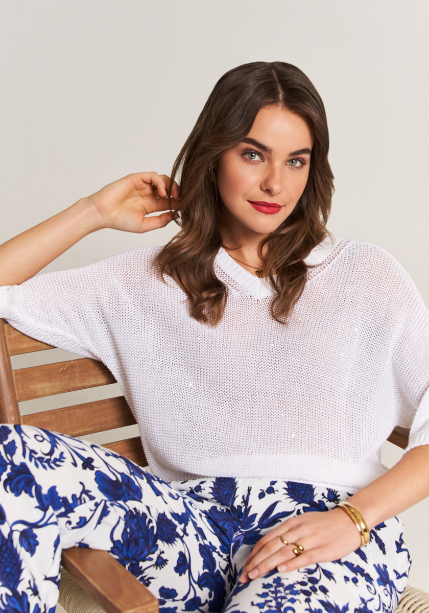 A woman with wavy brown hair and red lipstick sits on a wooden bench, wearing a loose white knit top and Princess Goes Hollywood blue floral-print elastic waist pants, accessorized with gold bracelets and rings.