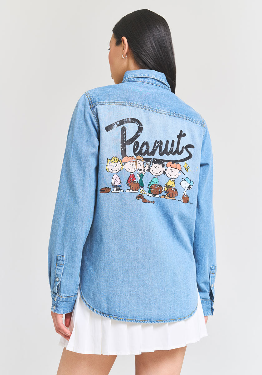 A woman with long dark hair, turned away to display the design, wears a light blue Princess Goes Hollywood denim shirt with Peanuts cartoon characters on the back and a short white skirt against a plain background.