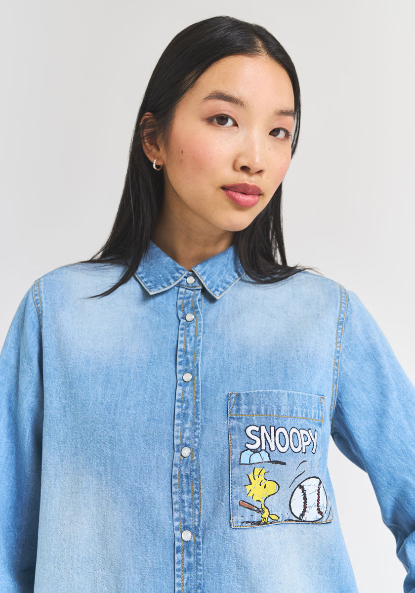 A young woman with straight black hair wears a light blue denim button-up shirt by Princess Goes Hollywood. The pocket shows Woodstock, a baseball, and “SNOOPY,” celebrating Peanuts 75th Anniversary. Background: plain white.
