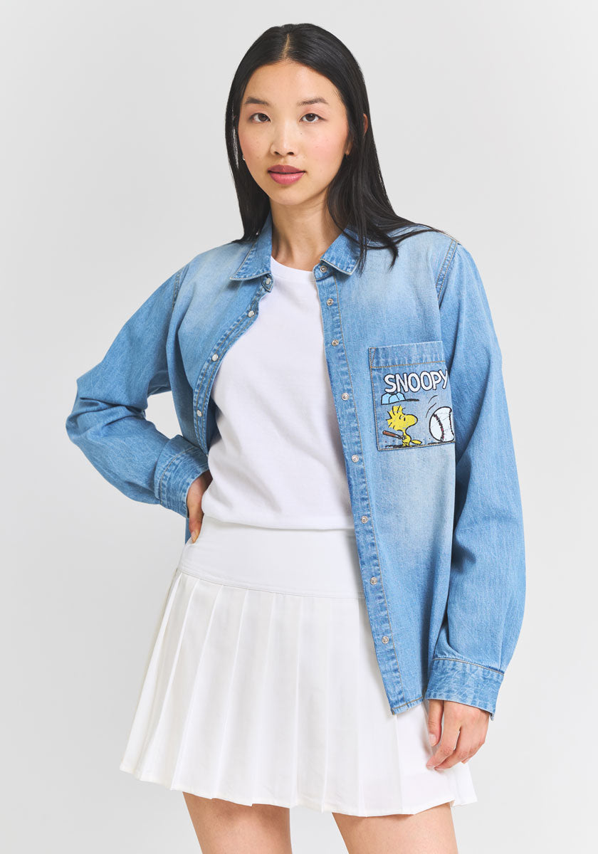A woman with long black hair wears a Princess Goes Hollywood light denim button-up shirt with a Snoopy &amp; Woodstock patch, paired with a white t-shirt and pleated skirt. She stands against a plain gray background, hand on her hip.