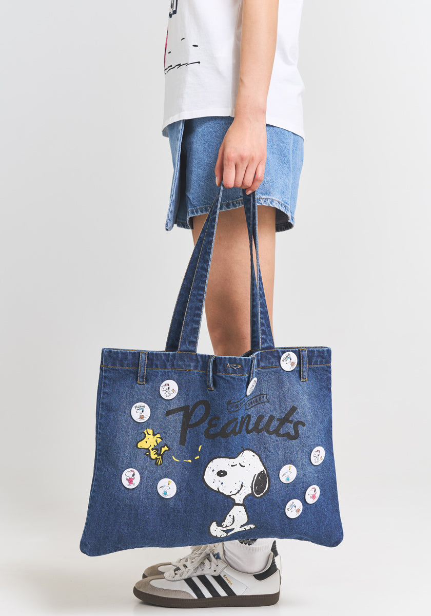 A person holds a blue Princess Goes Hollywood Denim-Shopper tote with the Peanuts 75th Anniversary Snoopy and Woodstock design, plus round character buttons. Theyre in a white tee, denim shorts, gray Adidas sneakers; background is plain white.