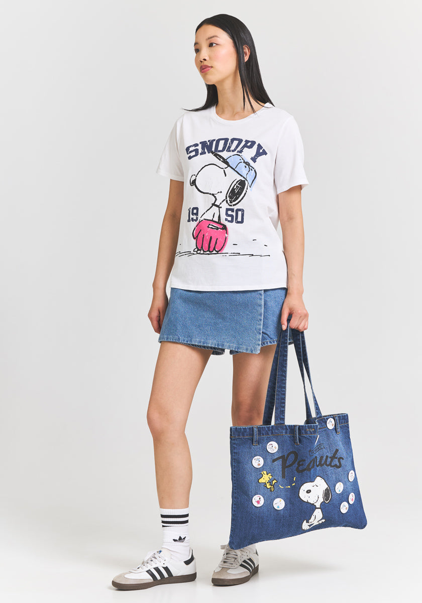 A woman with long straight hair wears a white Princess Goes Hollywood Snoopy T-shirt, a blue denim mini skirt, striped Adidas socks, and gray sneakers. She holds a Peanuts 75th Anniversary Denim-Shopper adorned with Snoopy, pins, and Peanuts.