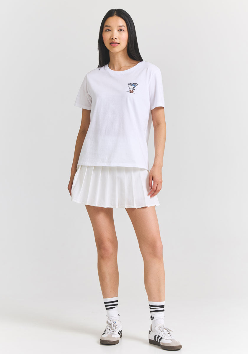 A woman with long black hair stands against a white background, wearing a Princess Goes Hollywood Snoopy T-shirt with a small chest graphic, paired with a white pleated tennis skirt, striped white socks, and white sneakers with black accents.