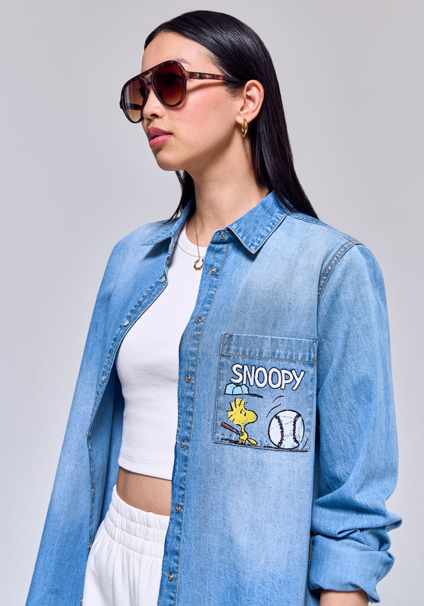 A woman with long dark hair wears brown sunglasses, gold hoops, a white crop top, and a Princess Goes Hollywood light blue denim shirt with a Snoopy-Spirit cartoon pocket, paired with white drawstring pants.