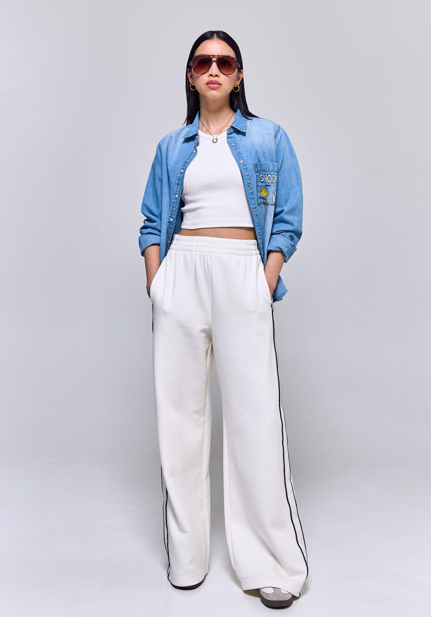 A woman with long dark hair poses against a light gray background wearing Princess Goes Hollywood: a blue denim shirt over a white crop top, loose white pants with black side stripes, gray sneakers, and large sunglasses. Her hands are in her pockets.