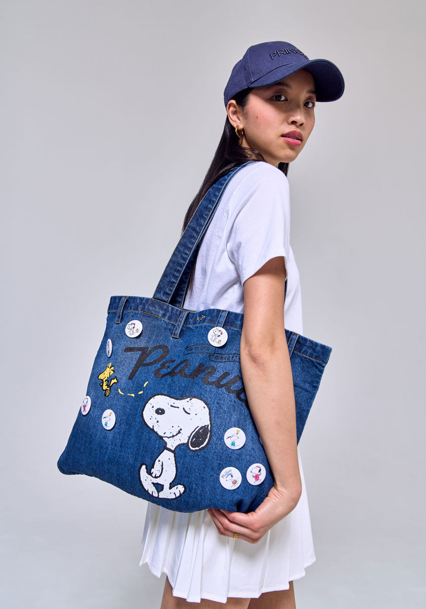 A woman in a white T-shirt, white pleated skirt, and navy cap stands in profile with a Princess Goes Hollywood Peanuts 75th Anniversary Denim-Shopper tote featuring Snoopy, Woodstock, “Peanuts” text, and colorful buttons.