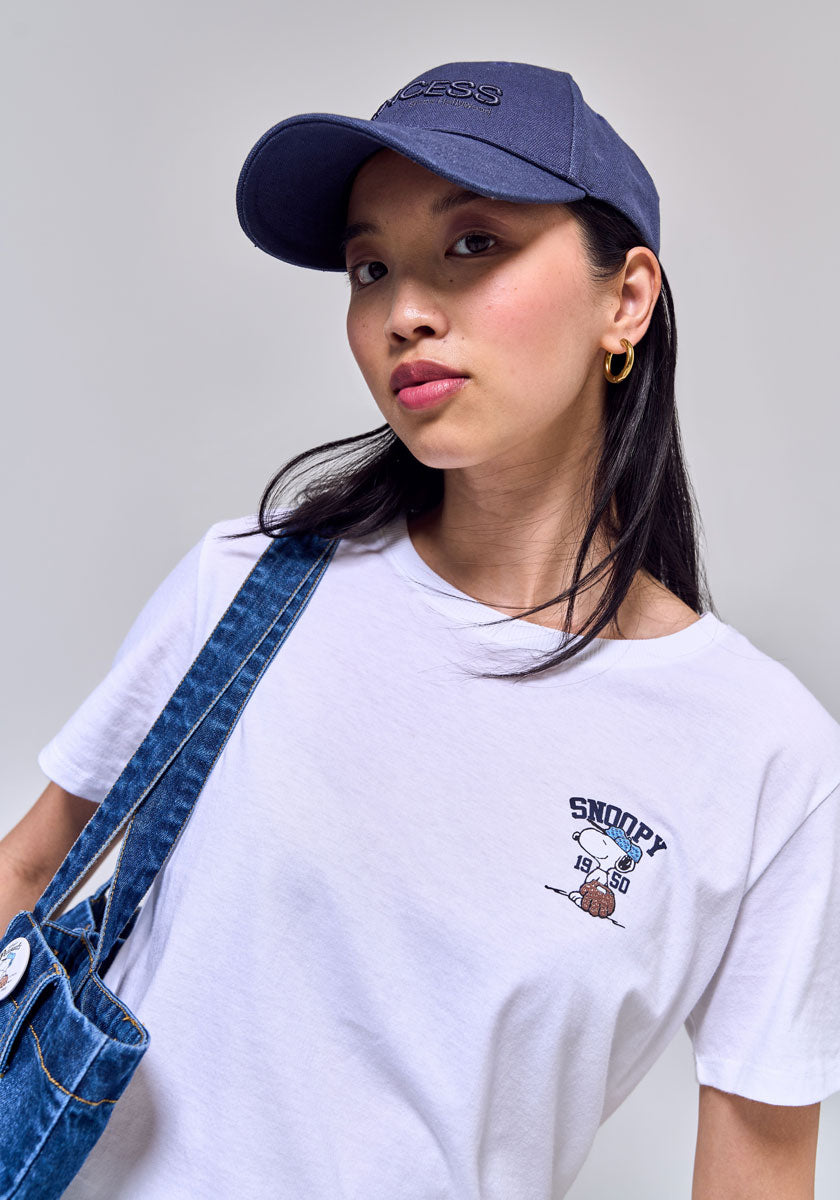 A young woman looks confidently at the camera, wearing a Princess Goes Hollywood white T-shirt with a small Snoopy design on the chest, a navy blue baseball cap, gold hoop earrings, and a denim strap over one shoulder. The background is plain light gray.