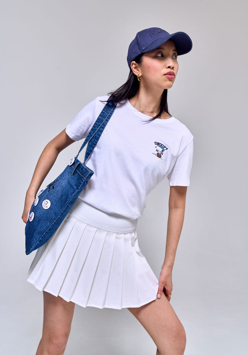 A woman poses against a plain background, wearing a white t-shirt, white pleated skirt, a blue baseball cap, and carrying a Princess Goes Hollywood Peanuts 75th Anniversary denim shopper decorated with pins and buttons.