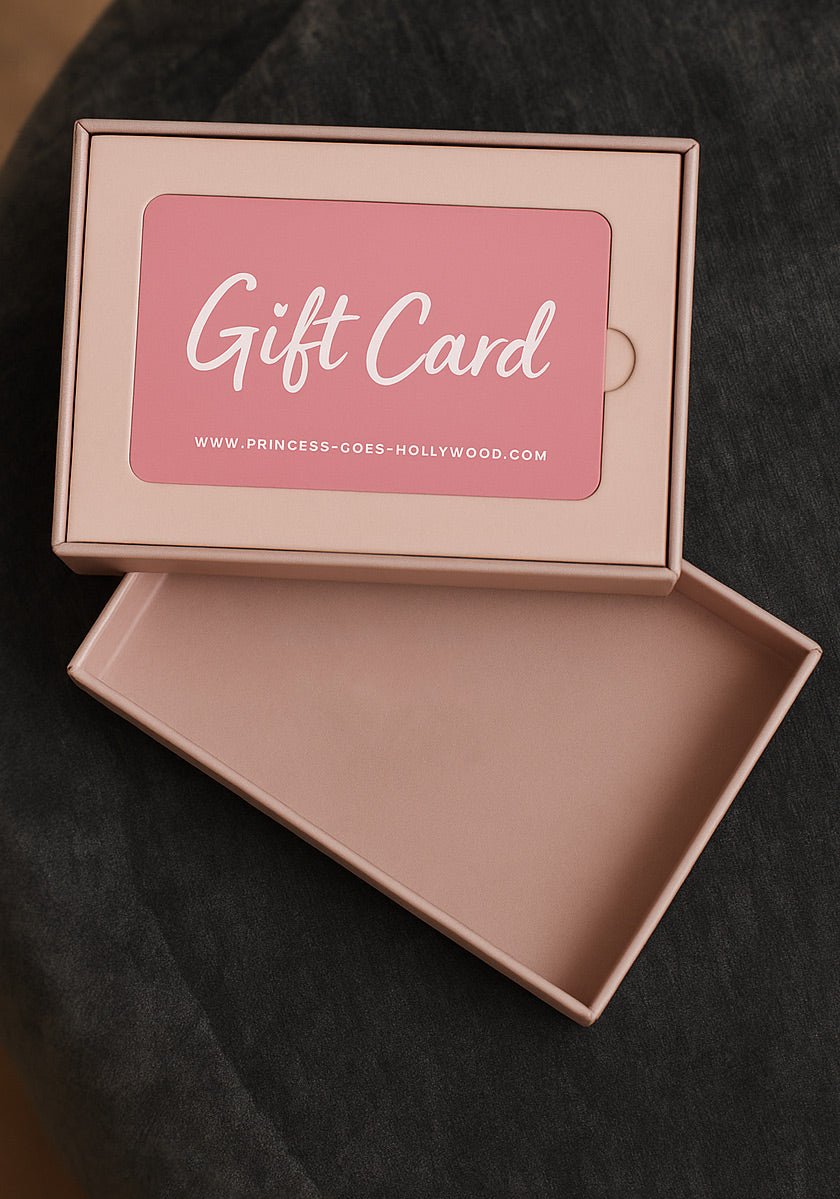 Eine rosafarbene Princess Goes Hollywood Geschenkkarte mit weißem &quot;Gift Card&quot;-Text und der Website ruht in einer passenden rosafarbenen Box, deren Deckel daneben auf einem dunklen Stoffhintergrund liegt - perfekt für jeden Mode-Liebhaber.