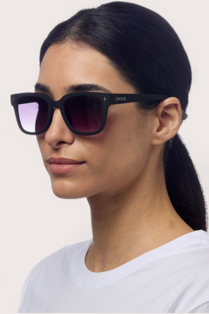 Eine Frau mit glattem, dunklem Haar in einem niedrigen Pferdeschwanz trägt eine schwarze Okkia-Sonnenbrille mit quadratischem Rahmen und Verlaufsgläsern, die den Markennamen auf dem Bügel trägt; sie blickt leicht zur Seite vor einem hellen Hintergrund.