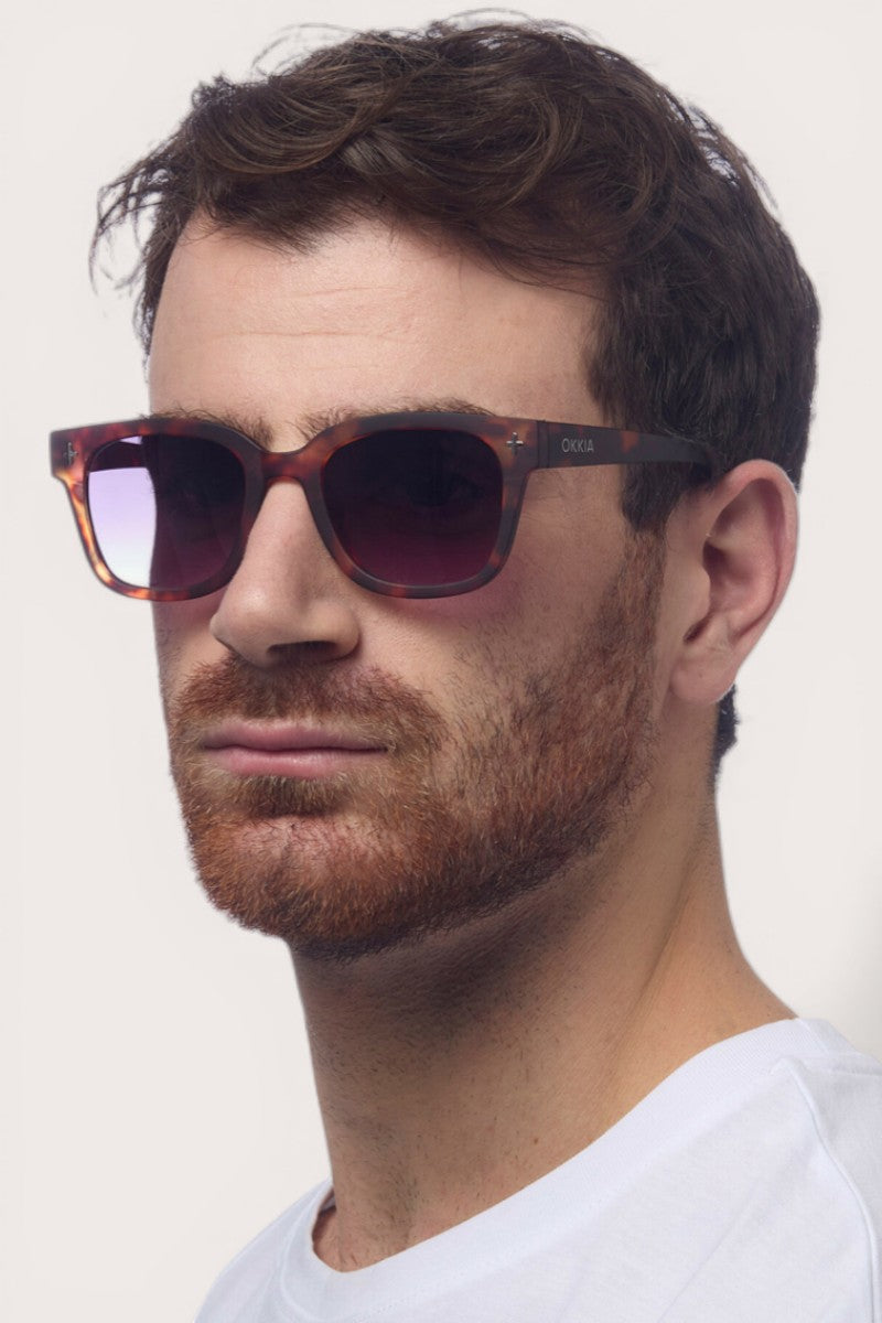 Ein Mann mit kurzen braunen Haaren, gestutztem rötlich-braunem Bart und ernstem Gesichtsausdruck trägt eine Okkia Sonnenbrille mit quadratischem Rahmen und UV 400 Schutz und ein weißes Hemd mit Rundhalsausschnitt vor einem schlichten hellen Hintergrund.