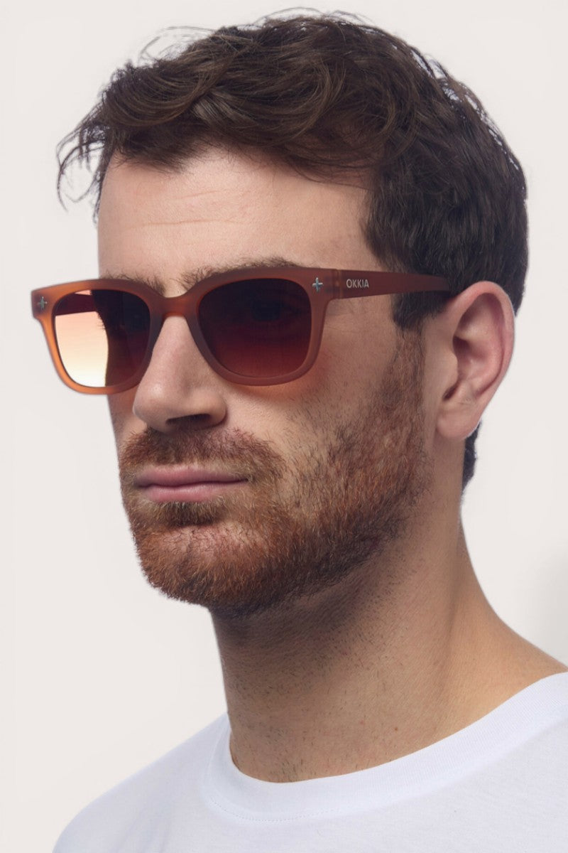 Ein Mann mit kurzen braunen Haaren und gestutztem Bart trägt eine braune Sonnenbrille mit Farbverlauf von Okkia (UV400, quadratischer Rahmen) und ein weißes T-Shirt, wobei er vor einem einfarbigen hellen Hintergrund leicht nach rechts blickt. Auf dem Bügel der Sonnenbrille ist das Wort OKKIA zu lesen.