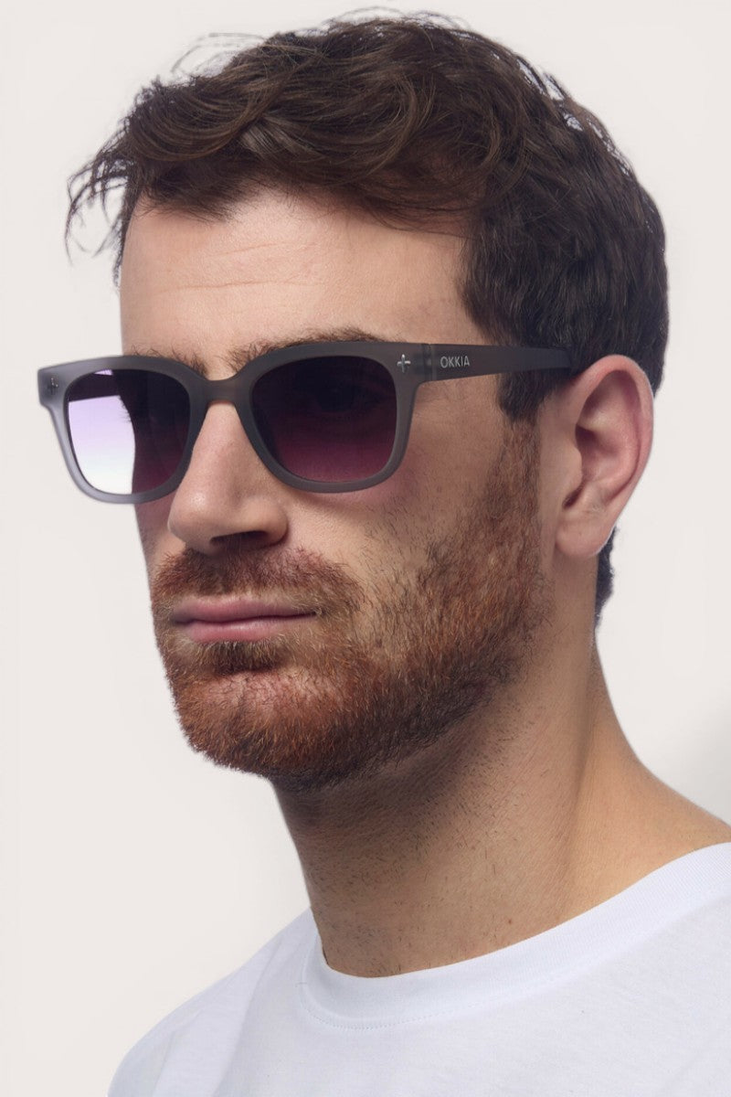 Ein Mann mit kurzen braunen Haaren und gestutztem Bart trägt eine große, dunkle Okkia-Sonnenbrille mit UV-400-Schutz und Square-Frame-Design. Er blickt leicht nach rechts vor einem schlichten, hellen Hintergrund, sein Ausdruck ist neutral.