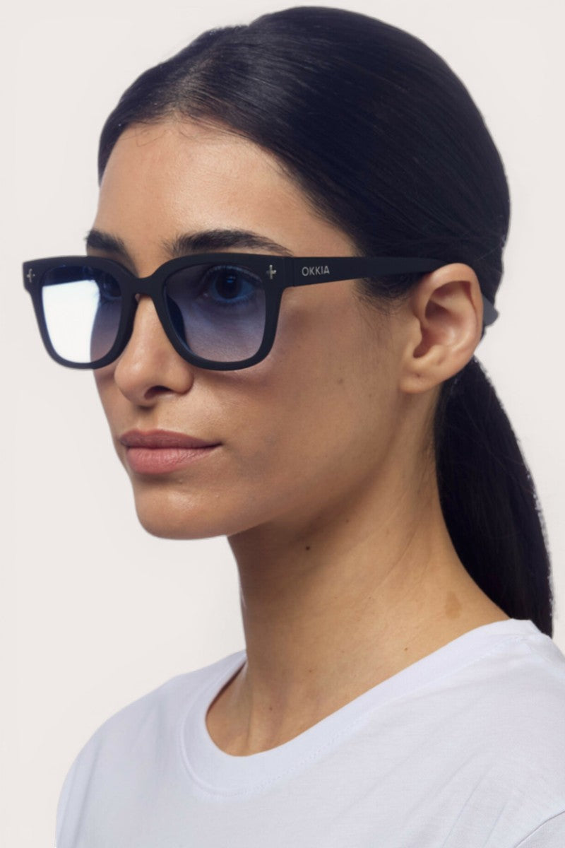 Eine Frau mit glattem dunklem Haar in einem niedrigen Pferdeschwanz trägt eine schwarze Okkia-Sonnenbrille mit Verlaufsgläsern und einem quadratischen Rahmendesign. Sie hat einen neutralen Gesichtsausdruck und trägt ein schlichtes weißes Oberteil vor einem hellen Hintergrund.