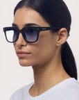 Eine Frau mit glattem dunklem Haar in einem niedrigen Pferdeschwanz trägt eine schwarze Okkia-Sonnenbrille mit Verlaufsgläsern und einem quadratischen Rahmendesign. Sie hat einen neutralen Gesichtsausdruck und trägt ein schlichtes weißes Oberteil vor einem hellen Hintergrund.