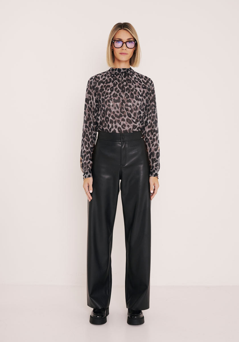 Eine Person mit glattem blondem Haar und Brille steht vor einem weißen Hintergrund, trägt eine Princess Goes Hollywood-Bluse mit Wildleopardenmuster, die in eine schwarze Lederhose mit weitem Bein und schwarzen Plateauschuhen gesteckt ist, und blickt neutral nach vorn.