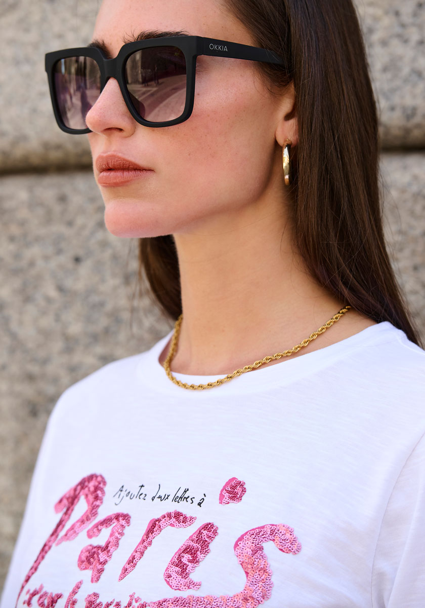 Eine Frau in übergroßer Okkia-Sonnenbrille mit UV-400-Schutz, goldenen Ohrringen und einer goldenen Kettenhalskette steht an einer Steinmauer. Sie hat glattes braunes Haar und trägt ein weißes T-Shirt mit rosa Glitzeraufdruck "Paris".