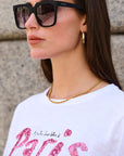Eine Frau in übergroßer Okkia-Sonnenbrille mit UV-400-Schutz, goldenen Ohrringen und einer goldenen Kettenhalskette steht an einer Steinmauer. Sie hat glattes braunes Haar und trägt ein weißes T-Shirt mit rosa Glitzeraufdruck "Paris".