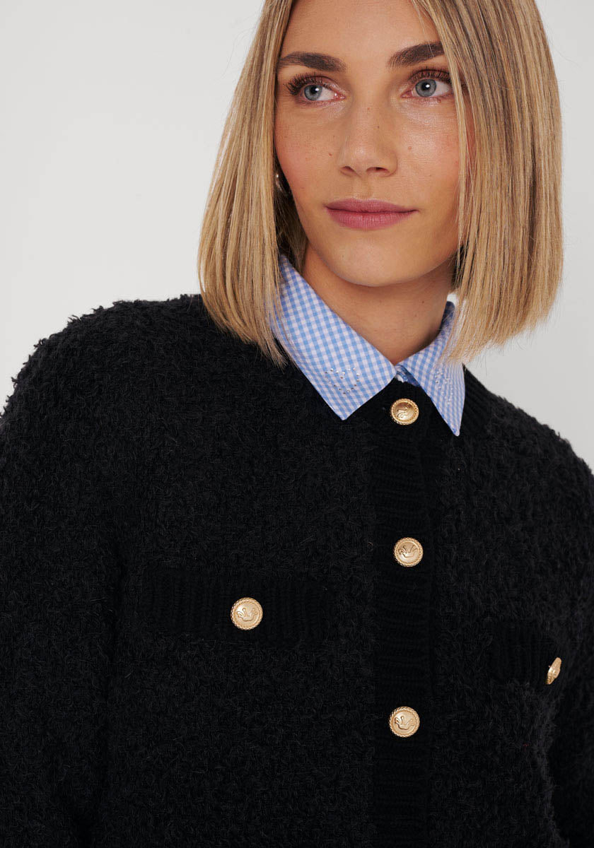 Eine Frau mit einem Bob-Haarschnitt und hellen Augen trägt eine strukturierte Princess Goes Hollywood Strickjacke aus Wolle-Alpaka-Mix mit goldenen Knöpfen über einem blau-weiß karierten Hemd mit Kragen und blickt zur Seite vor einem schlichten hellen Hintergrund.