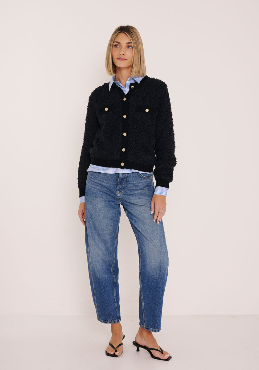 Eine Frau mit glattem blondem Haar posiert entspannt. Sie trägt eine Princess Goes Hollywood schwarze Strickjacke aus Wolle-Alpaka-Mix mit goldfarbenen Knöpfen über einem hellblauen Hemd, lockeren Jeans und schwarzen Heels vor weißem Hintergrund.