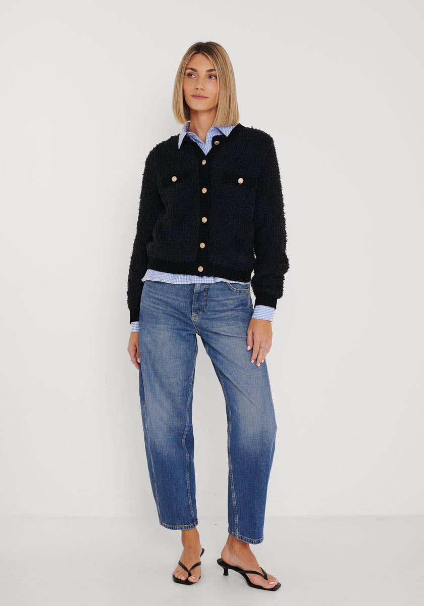 Eine Frau trägt eine schwarze Strickjacke aus Wolle-Alpaka-Mix mit goldfarbenen Knöpfen von Princess Goes Hollywood über einem hellblauen Hemd, relaxten Jeans und schwarzen Heels. Ihr blondes Haar ist schulterlang, ihr Blick neutral.