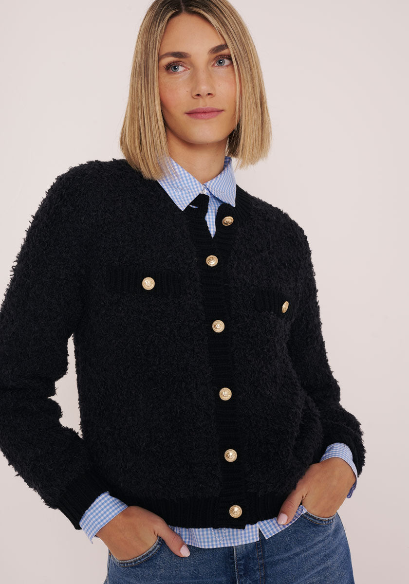 Eine Frau mit glattem blondem Haar trägt eine Princess Goes Hollywood-Strickjacke aus einem Woll-Alpaka-Gemisch mit großen goldenen Knöpfen über einem blau-weiß-karierten Hemd. Sie steht mit den Händen in den Taschen ihrer Jeans vor einem hellen Hintergrund und blickt zur Seite.