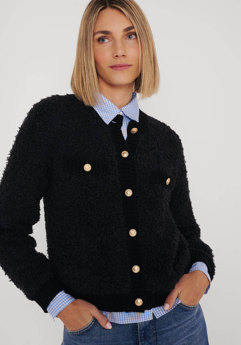 Eine Frau mit glattem, schulterlangem blondem Haar trägt eine Strickjacke aus Wolle-Alpaka-Mix mit goldfarbenen Knöpfen von Princess Goes Hollywood über einem blauen Gingham-Hemd, steht vor hellem Hintergrund und lächelt leicht.