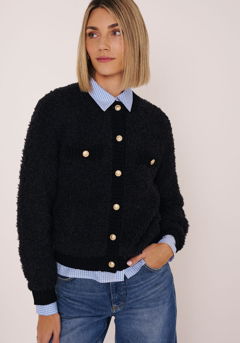 Eine Frau mit blondem Bob trägt eine Princess Goes Hollywood Strickjacke aus Wolle-Alpaka-Mix mit goldfarbenen Knöpfen über einem blau karierten Hemd. Sie steht vor einem hellen Hintergrund, eine Hand in der Jeanstasche, und wirkt entspannt.