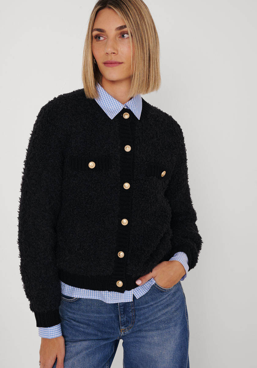 Eine Frau mit glattem blondem Haar trägt eine Princess Goes Hollywood schwarze Strickjacke aus Wolle-Alpaka-Mix mit goldfarbenen Knöpfen über einem blau-weiß karierten Hemd, Jeans und blickt nach links vor weißem Hintergrund.