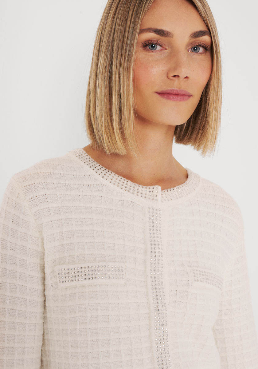 Eine Frau mit glattem, schulterlangem blondem Haar trägt eine cremefarbene Princess Goes Hollywood-Strickjacke aus Baumwolle und Kaschmir mit Gittermuster, Ziertaschen und Strassperlenbesatz. Sie blickt nach vorne und lächelt leicht vor einem weißen Hintergrund.