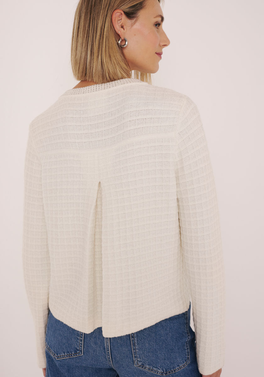 Eine Frau mit glattem blondem Haar und silbernen Ohrringen ist von hinten zu sehen. Sie trägt eine naturfarbene Baumwolle-Kaschmir-Mix Strickjacke mit Kellerfalte und blaue Jeans vor einem schlichten hellen Hintergrund.