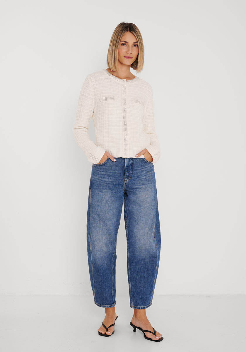 Eine Frau mit glattem, schulterlangem blondem Haar posiert selbstbewusst vor einer weißen Wand. Sie trägt eine hellbeige Baumwolle-Kaschmir-Mix Strickjacke von Princess Goes Hollywood, weite blaue High-Waisted-Jeans und schwarze hochhackige Sandalen.