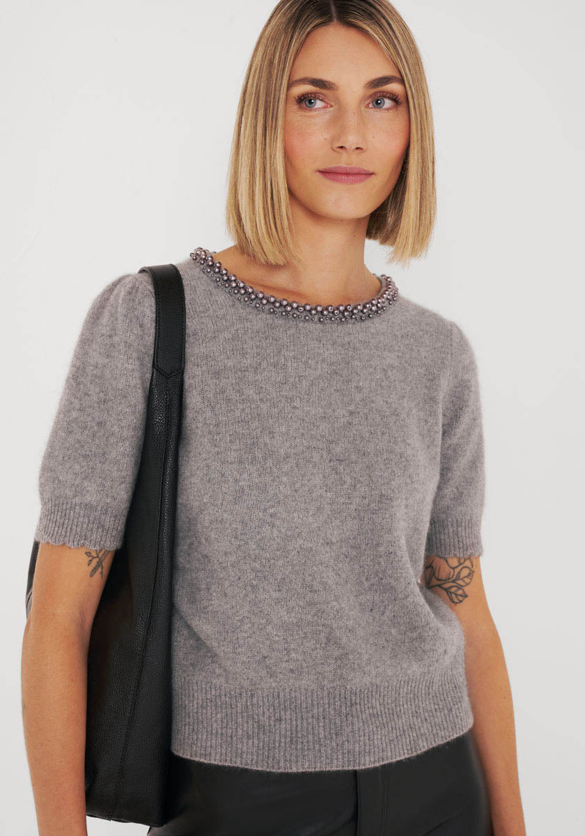 Eine Frau mit glattem, schulterlangem blondem Haar trägt einen weichen, hellgrauen Strickpullover von Princess Goes Hollywood mit einem perlenbesetzten Ausschnitt. Sie trägt eine schwarze Tasche und zeigt Unterarmtattoos vor einem schlichten weißen Hintergrund.