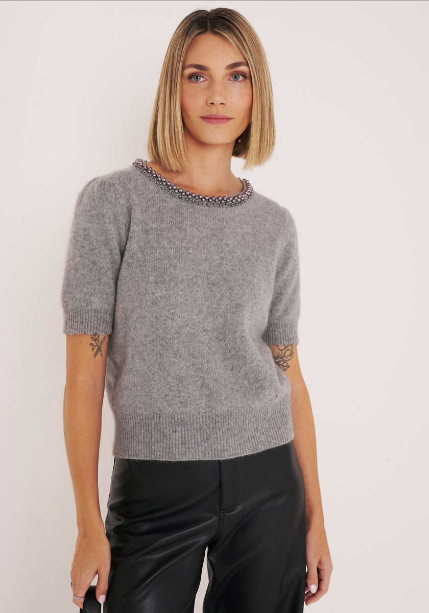 Eine Frau mit glattem blondem Bob trägt einen grauen Princess Goes Hollywood Strickpullover mit perlenbesetztem Ausschnitt und eine schwarze Lederhose. Sie steht vor einem weißen Hintergrund und blickt mit entspanntem Blick nach vorn.