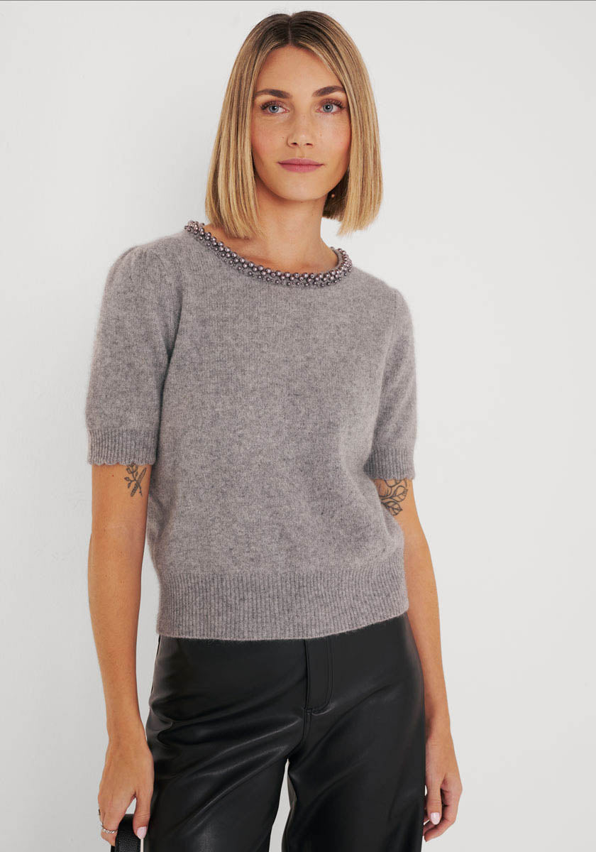 Eine Frau mit glattem, schulterlangem blondem Haar trägt einen hellgrauen Princess Goes Hollywood Strickpullover aus Wolle-Waschbärfaser-Mix mit Perlen am Ausschnitt und eine schwarze Lederhose. Sie steht vor einem schlichten weißen Hintergrund und hat einen neutralen Gesichtsausdruck.