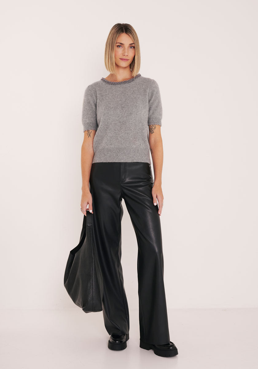 A woman with Bob trägt einen grauen Strickpullover mit Perlen am Kragen, eine schwarze weite Lederhose und Schuhe von Princess Goes Hollywood. Vor weißem Hintergrund hält sie eine große schwarze Tasche und blickt in die Kamera.