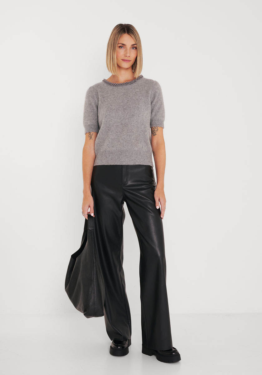 Eine Frau mit Bob trägt einen kurzärmeligen grauen Strickpullover und eine schwarze Lederhose mit weitem Bein von Princess Goes Hollywood und hält eine große schwarze Tasche in ihrer linken Hand. Sie steht vor einer schlichten weißen Wand, trägt schwarze Schuhe und einen neutralen Gesichtsausdruck.