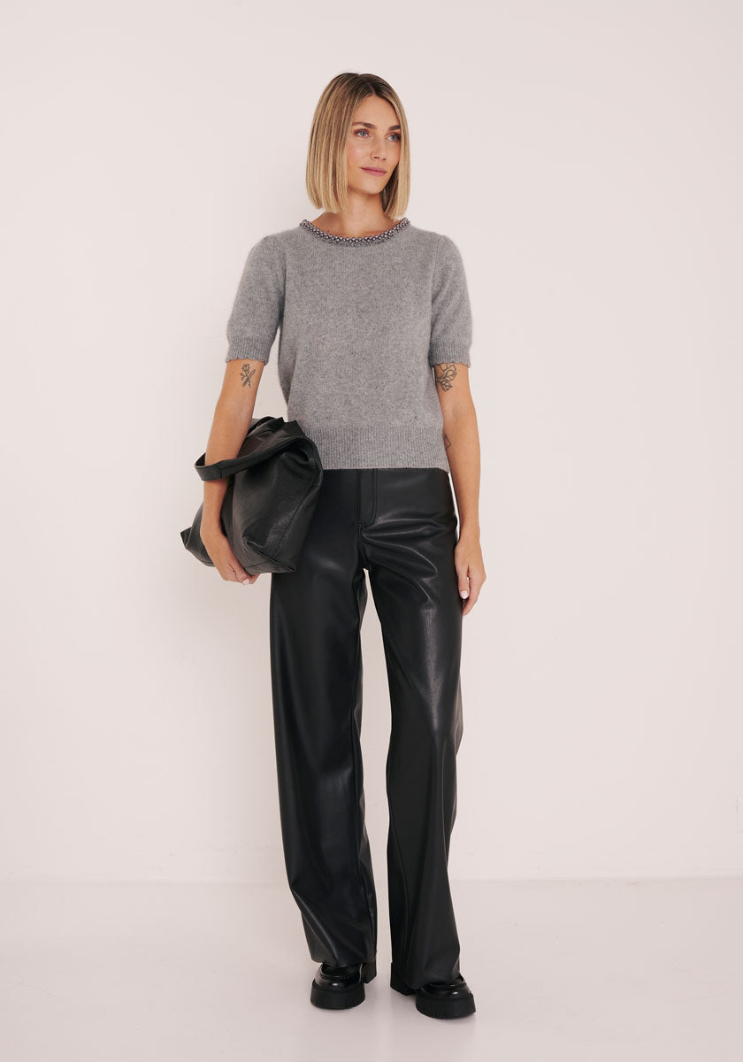 Eine Frau mit einem Bob-Haarschnitt steht vor einem weißen Hintergrund und trägt einen grauen Strickpullover von Princess Goes Hollywood, eine schwarze Lederhose und schwarze Schuhe. Sie hält eine große schwarze Clutch, ihre tätowierten Arme sind sichtbar, während sie zur Seite lächelt.