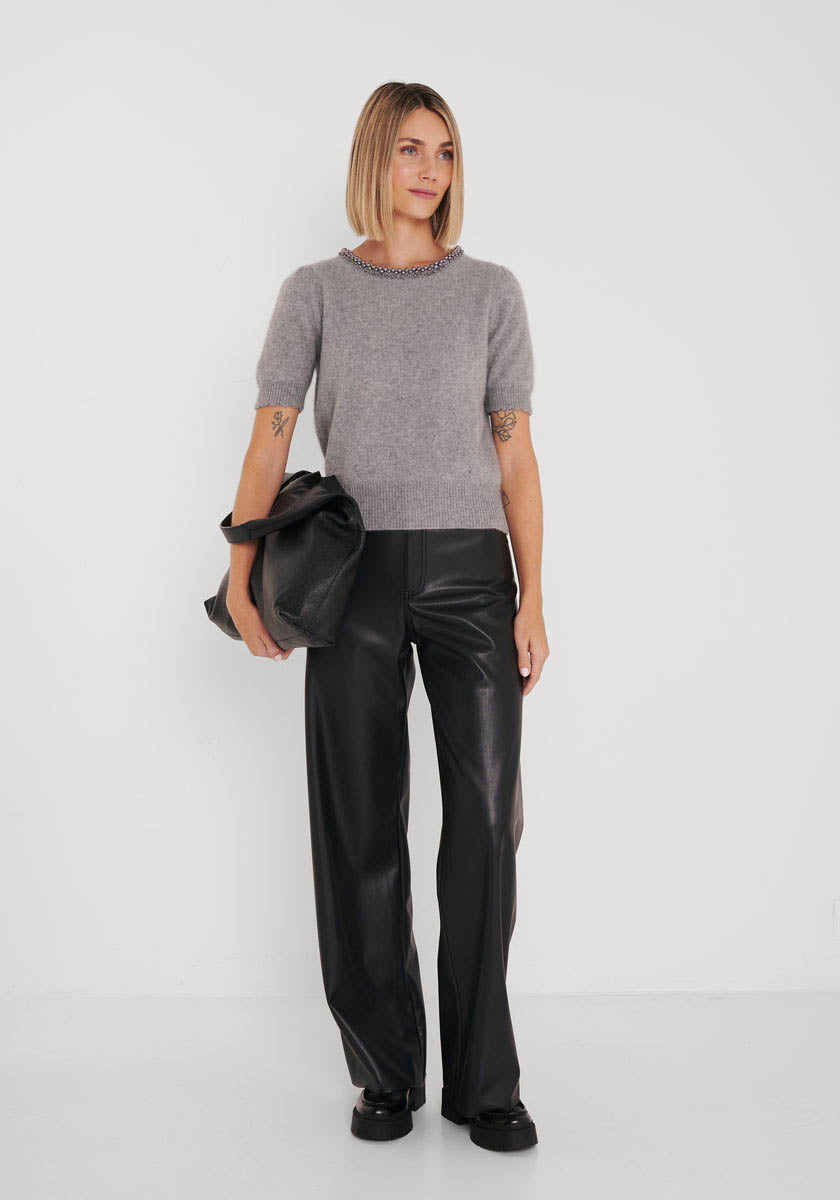 Eine Frau mit glattem, schulterlangem blondem Haar steht vor einer weißen Wand und trägt den kurzärmeligen Strickpullover von Princess Goes Hollywood im Wolle-Waschbärfaser-Mix, eine schwarze Lederhose mit weitem Bein, schwarze Schuhe und eine große schwarze Tasche.