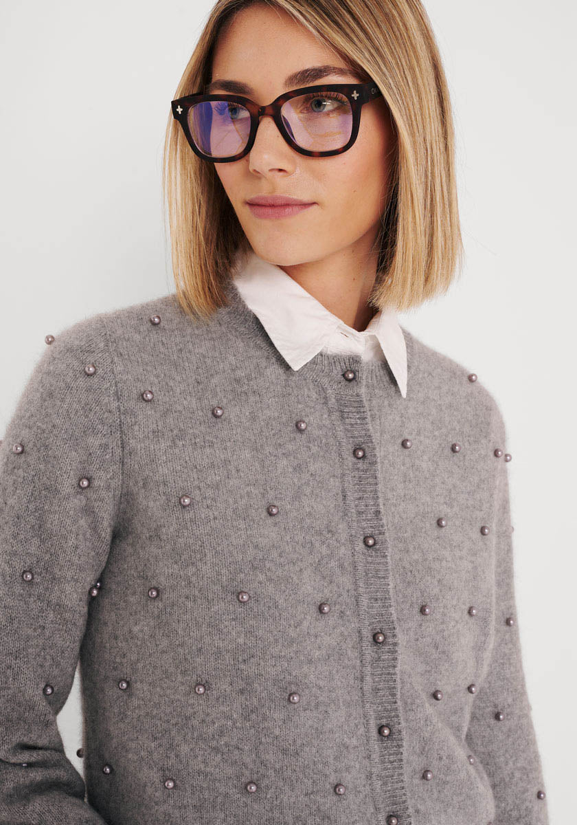 Eine Frau mit geradem blonden Haarschnitt, einer Schildpattbrille und einem weißen Hemd mit Kragen trägt eine graue Strickjacke aus Wollgemisch mit Perlenknöpfen von Princess Goes Hollywood. Sie blickt leicht nach rechts und wirkt ruhig vor einem schlichten Hintergrund.