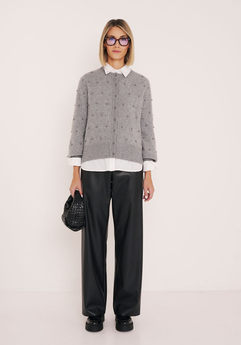 Eine Person mit kurzen blonden Haaren und Brille steht vor einer schlichten hellen Wand. Sie trägt eine graue Princess Goes Hollywood Strickjacke mit Perlenknöpfen über einem weißen Hemd mit Kragen, eine schwarze Hose mit weitem Bein, Plateauschuhe und hält eine kleine schwarze Handtasche in der Hand.