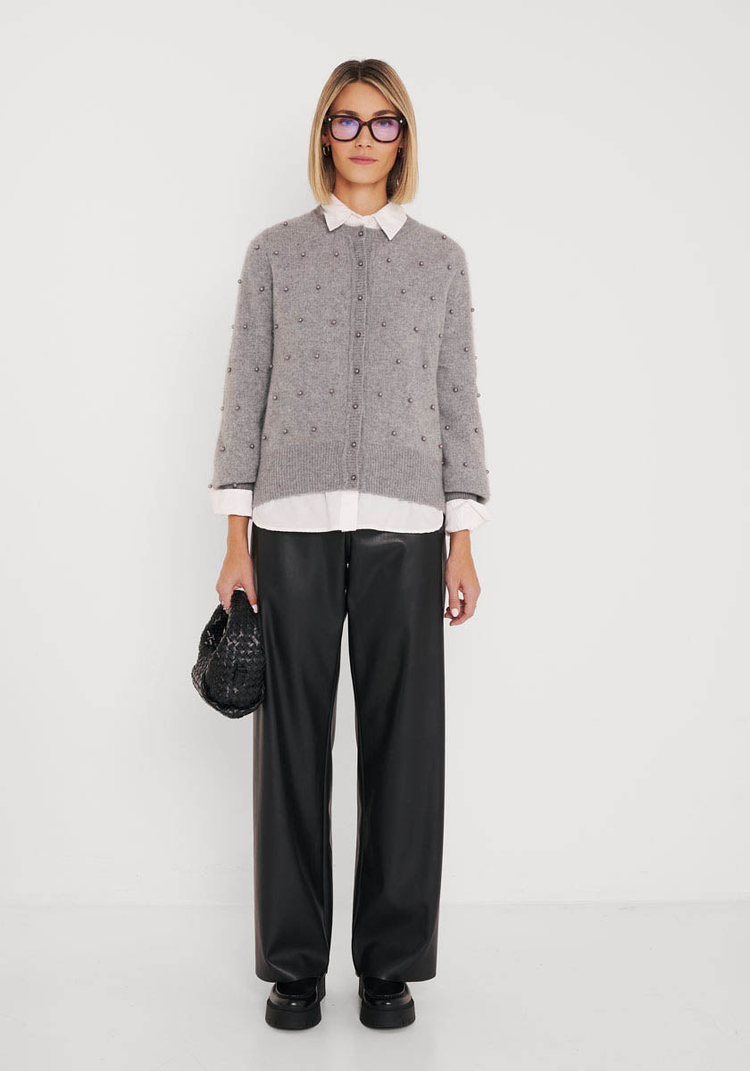 Eine Person mit blondem Haarschopf und lila Brille trägt eine graue Princess Goes Hollywood Wollmix Strickjacke mit Perlenknöpfen über einem weißen Hemd mit Kragen, eine schwarze Lederhose und Plateauschuhe und hält eine schwarze Handtasche vor einem weißen Hintergrund.